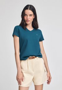 Tealfärgad t-shirt med v-ringning och korta ärmar, gjord av mjukt tyg. Kombination med ljusbeige shorts och ett brunt bälte. Minimalistisk design.