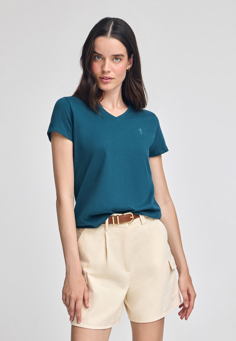 Tealfärgad t-shirt med v-ringning och korta ärmar, gjord av mjukt tyg. Kombination med ljusbeige shorts och ett brunt bälte. Minimalistisk design.