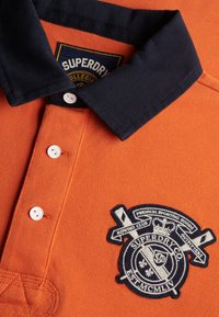 Polo orange avec col noir, boutons blancs et un écusson brodé du club d'aviron Superdry noir et blanc sur la poitrine.