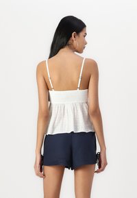 Balta smockinė palaidinė su plonomis petnešomis, tekstūruota medžiaga ir platėjantis kraštas, derinama su tamsiai mėlynomis šortais, turinčiais rišamųjų detalių.