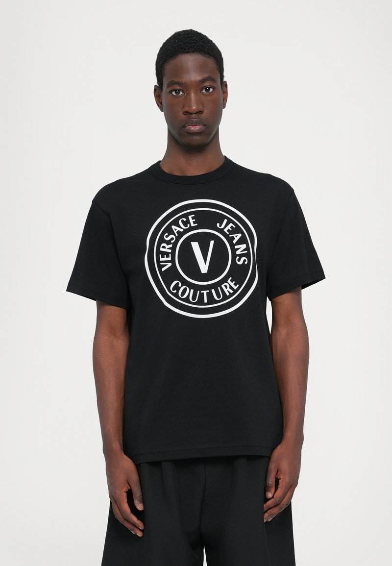 Svart t-skjorte med korte ermer med et stort rundt hvitt logo som har teksten "VERSACE JEANS COUTURE" og et sentralt "V"-symbol.