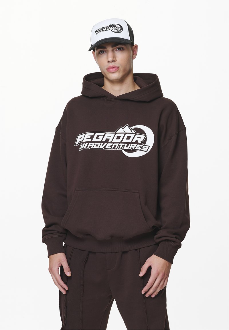 Brauner Hoodie mit einem großen weißen "Pegador Adventures"-Logo, Kängurutasche und gerippten Bündchen, kombiniert mit einer weiß-schwarzen Mütze.