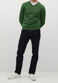 Homme portant un pull vert à col en V, un jean bleu foncé et des baskets blanches, debout les mains derrière le dos devant un fond uni.