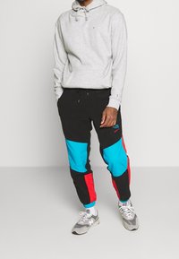 Sudadera gris con bolsillo frontal, combinada con pantalones de chándal negros que tienen paneles en turquesa y rojo. Llevada con zapatillas grises.