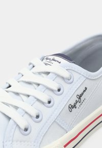 Light blue canvas sneaker with white laces, rubber sole, and red and navy stripes. Features branded label on the tongue.