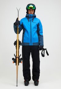 Skibekleidung mit einer blau-grauen wasserdichten Jacke und schwarzen Hosen. Die Person hält gelb-schwarze Skier und trägt einen Helm sowie eine Skibrille.