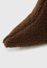 Material texturat maro, cu o suprafață moale și pufoasă, având o formă ascuțită și detalii cusute de-a lungul marginii.