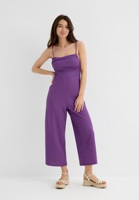 Jumpsuit morado de canalé con un torso ajustado, silueta de pierna ancha y tirantes finos ajustables. Llevado con sandalias de plataforma en un tono beige claro.