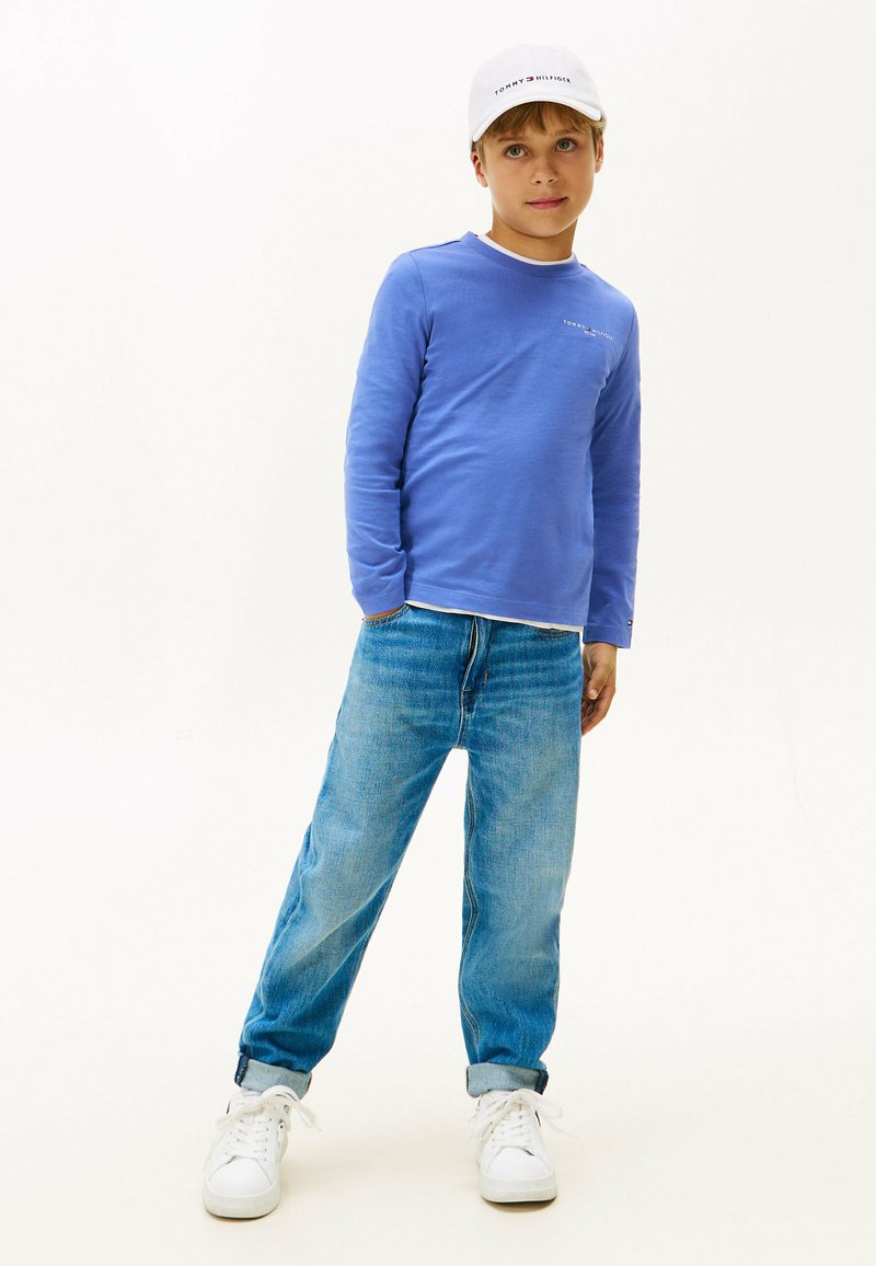 Blaue Langarmbluse mit einem weißen Kragen, kombiniert mit hellblauen Jeans und weißen Sneakers, ergänzt durch eine weiße Kappe mit Logo.