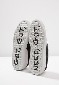 Mustat ja valkoiset tennarit, joissa on tekstuuripohjat ja kohotettuja pyramideja. Pohjissa näkyy isot mustat kirjaimet "GOT" ja "NEED".