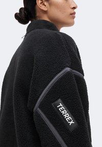 Personne ayant les cheveux en chignon portant une veste polaire noire texturée avec un patch TERREX sur le haut de la manche.