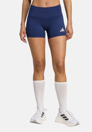 Dunkelblaue Sportshorts mit hohem Taillenbund, ausgestattet mit einem weißen Adidas-Logo. Getragen mit knielangen weißen Socken und weißen Turnschuhen.