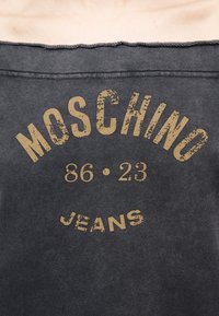 Σκούρο γκρι ύφασμα με φθαρμένο μπεζ κείμενο που γράφει «MOSCHINO 86 · 23 JEANS» με έντονα γράμματα.