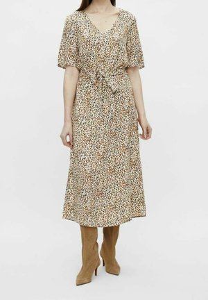 Kobieta w beżowej i brązowej sukience midi w cętki, z krótkimi rękawami, dekoltem w serek, wiązaną talią i beżowymi zamszowymi botkami na wysokim obcasie.
