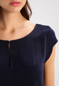 Žena v námořnické modré blůze s krátkým rukávem a kulatým výstřihem, se dvěma svislými otvory ve tvaru klíče v přední části.