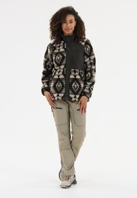 Veste en polaire avec un motif tribal noir et beige, fermeture éclair à l'avant et col haut. Associée à un pantalon kaki à zip et des chaussures de randonnée.