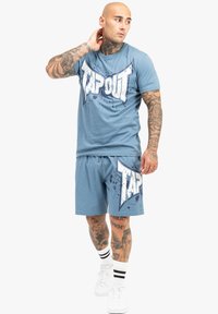 Mėlynos medvilninės marškinėlių ir šortų komplektas, kuriame puikuojasi didelis baltas "TAPOUT" grafikas su purškimo akcentais, derinamas su baltais sportiniais bateliais ir dryžuotomis kojinėmis.