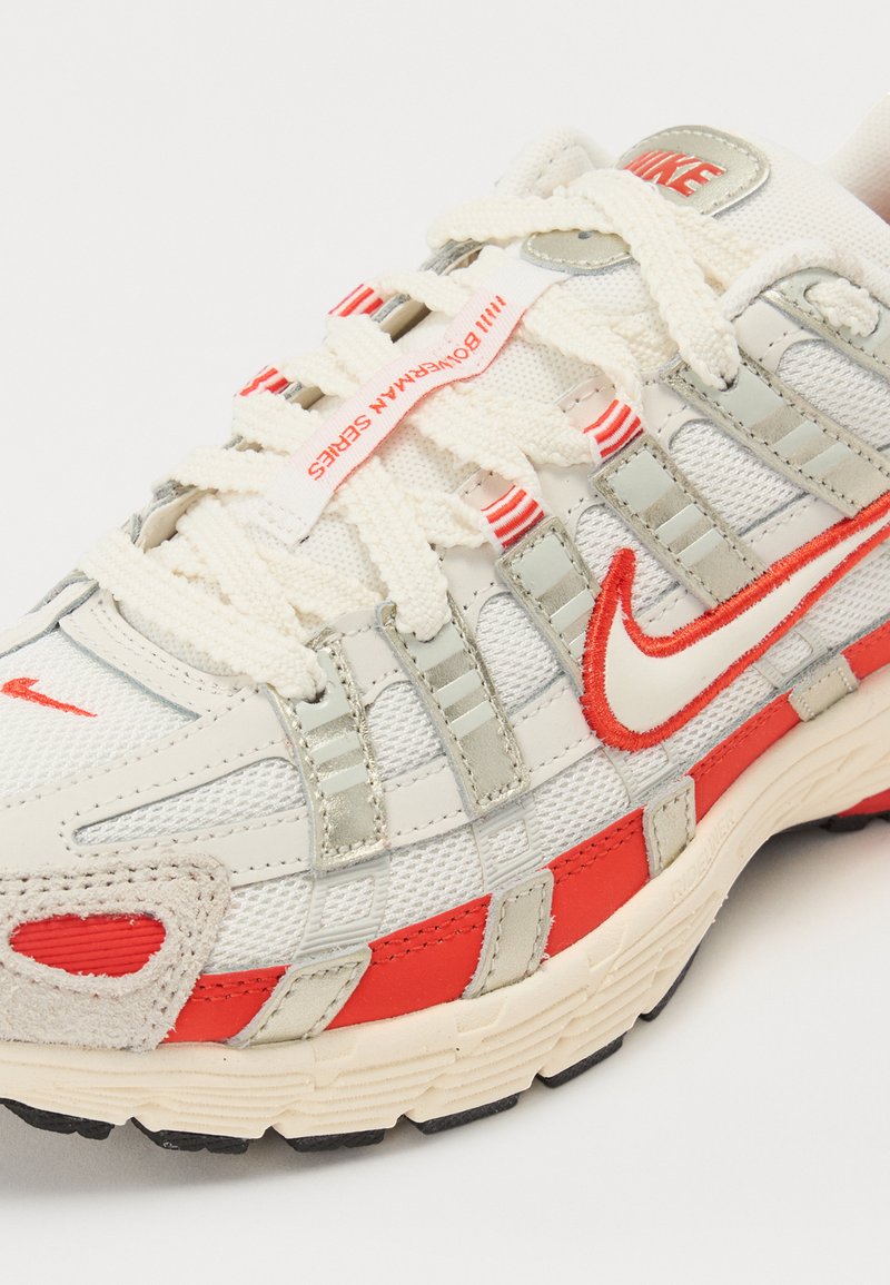 Nike Laufschuh mit einem Mesh-Obermaterial in Creme und Grau. Verfügt über rote Akzente, eine strukturierte Gummisohle und flache Schnürsenkel für einen sicheren Sitz.