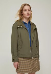 Giacca leggera verde oliva con cappuccio, chiusura con zip, due tasche frontali e polsini regolabili, abbinata a una camicia di jeans blu e una gonna a scacchi.