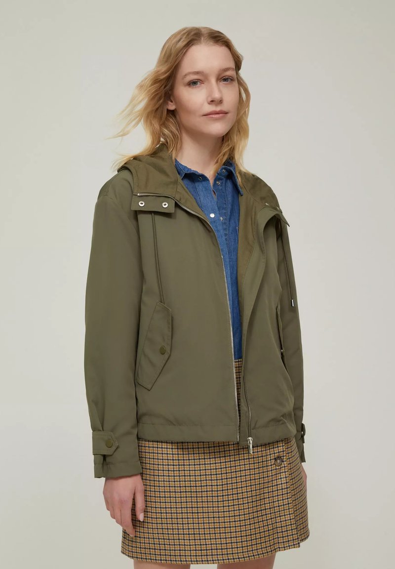Giacca leggera verde oliva con cappuccio, chiusura con zip, due tasche frontali e polsini regolabili, abbinata a una camicia di jeans blu e una gonna a scacchi.