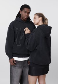 Sudaderas negras de ajuste holgado con mangas largas, con un diseño gráfico texturizado. La modelo las combina con pantalones grises claros y una minifalda negra.