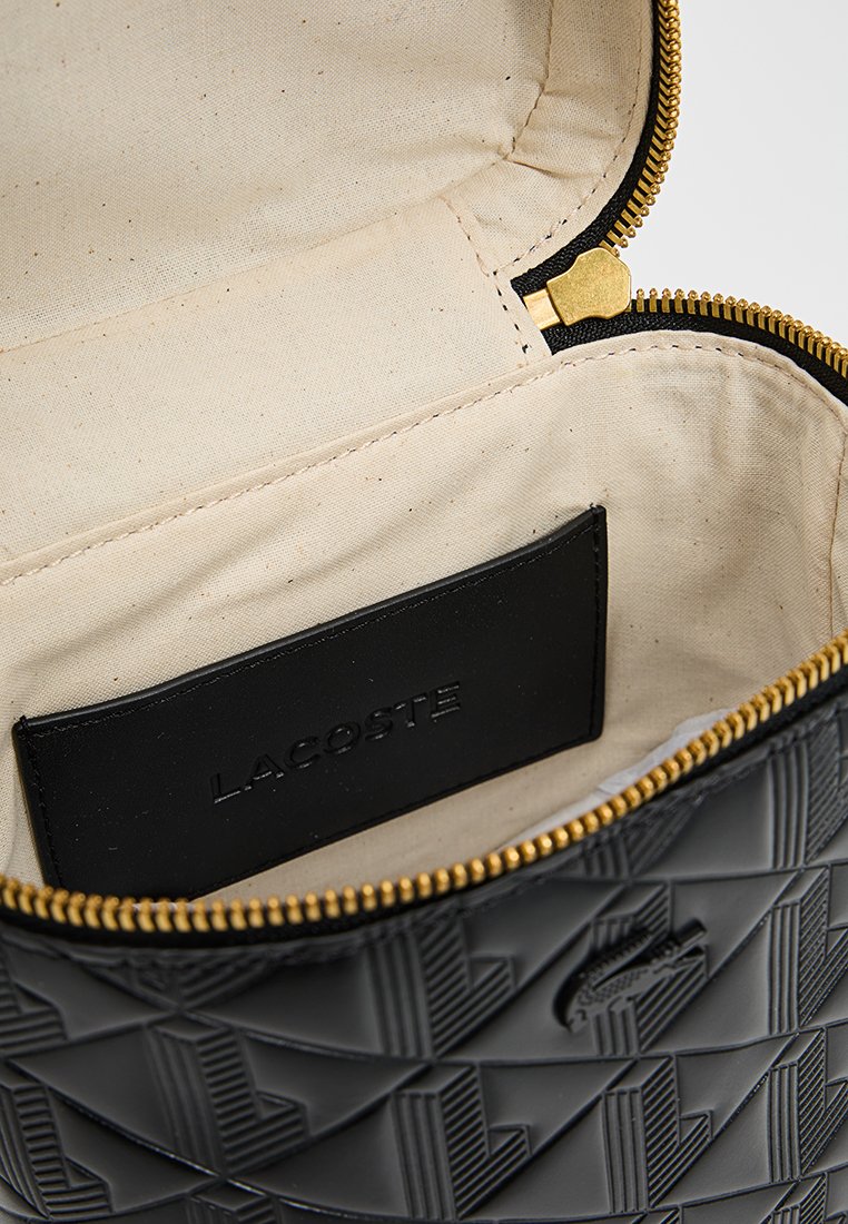 Sac fourre-tout noir à motifs avec fermeture éclair dorée et doublure beige à l'intérieur, comportant une étiquette en cuir noir avec "LACOSTE" en relief.