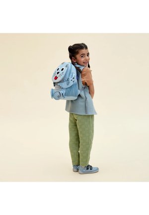 Lächendes Mädchen, das grüne Hose und ein braunes Hemd trägt, trägt einen blauen Rucksack mit einem verspielten Robbengesicht und einer Wasserflasche an der Seite.