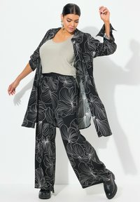 Zwart bloemetjesoutfit met een vloeiende lange jas met wijde mouwen, gecombineerd met losse broek en een lichtgrijze top. Zwarte chunky schoenen.