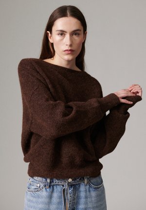 Femme aux cheveux bruns raides portant un pull marron ample et un jean bleu clair, posant les mains jointes devant un fond uni.