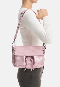 Sac à bandoulière en cuir rose avec une finition brillante, doté de deux poches avant, de détails à cordon et d'une bandoulière réglable cloutée.