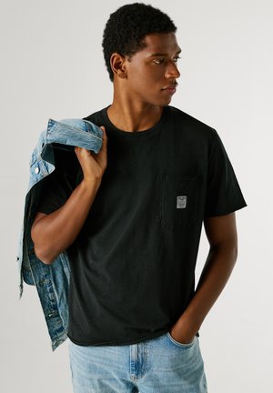 Zwart katoenen t-shirt met een borstzak, gecombineerd met een denim jas over de schouder; model draagt lichtblauwe jeans.