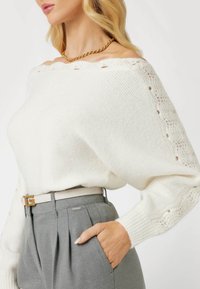 Maglione bianco a spalle scoperte in maglia con dettagli in pizzo. Abbinato a pantaloni grigi plissettati, con una cintura chiara dotata di una fibbia dorata.