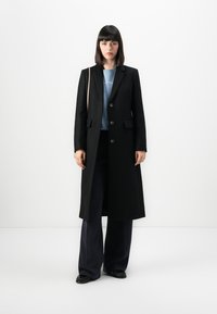 Donna in piedi che indossa un lungo cappotto nero, un maglione blu, pantaloni neri larghi, scarpe nere e porta una borsa a tracolla beige.