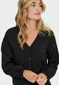 Čierna blúzka s výstrihom do V, gombíkmi po celej dĺžke a riasenými manžetami. Textúrovaná tkanina s voľným strihom a peplum detailom.