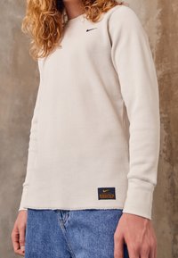 Long-sleeve, cream ribbed shirt with a black Nike logo on the chest and a label at the hem. Features fitted cuffs and a relaxed fit.