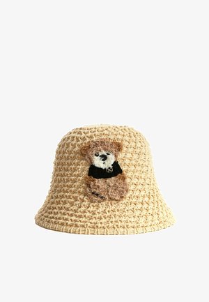 Sombrero de cubo beige de ganchillo con un pequeño parche de oso de peluche marrón que lleva una camisa negra en el frente.