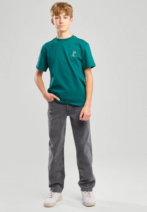 Teenager steht mit Hand in der Tasche, trägt grünes T-Shirt, graue Jeans und weiße Sneakers vor einfarbig grauem Hintergrund.