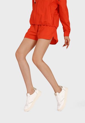 Persona che indossa pantaloncini e giacca arancione brillante, in posa a metà passo, con sneaker bianche con accenti beige su uno sfondo semplice.