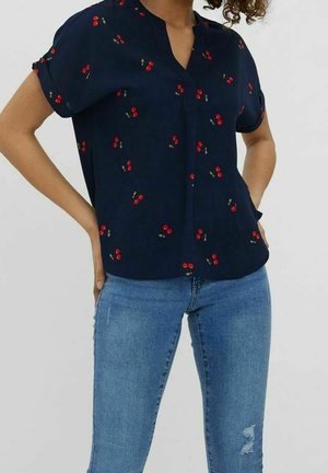 Kvinde iført en marineblå bluse med røde kirsebærmønstre og lyseblå, slidte skinny jeans, stående med hænderne på hofterne.