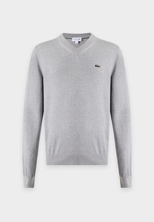 Jeune homme portant un pull gris à col en V par-dessus une chemise blanche, jean bleu, ceinture noire et baskets blanches, debout avec une main dans la poche.