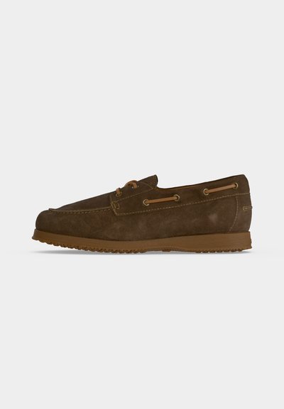 Mocassin en daim marron avec des détails en lacets de cuir et une semelle en caoutchouc sur un fond clair uni, vu de côté.