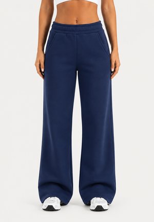 Femme portant un pantalon de jogging large bleu marine avec poches et des baskets blanches, vue de la taille jusqu’aux pieds sur un fond uni.