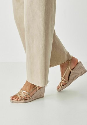 Gold strappy wedge sandals with a woven jute-style sole, featuring a textured upper and an open-toe design. Paired with light linen pants.