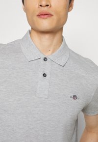 Homme portant un polo gris clair avec un petit logo GANT brodé sur la poitrine, deux boutons noirs fermés au col.
