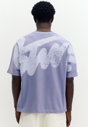 Homme portant un t-shirt violet ample avec un grand motif abstrait de taches de peinture blanches dans le dos, associé à un pantalon de couleur claire.