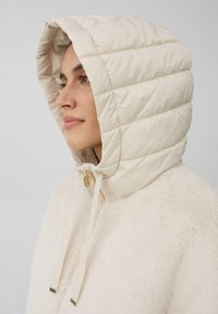 Gevoerde capuchonjas in crèmekleur met een geborduurde textuur. Heeft een harige faux fur body, gouden hardware en een verstelbare trekkoord bij de nek.