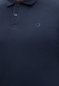 Μπλε ναυτικό πόλο με δύο κουμπιά και μικρό, τονισμένο κέντημα λογότυπου στο αριστερό στήθος.