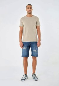 Bež majica s okruglim izrezom, plave traper šortse s istrošenim detaljima i sive tenisice s bijelim naglascima, na muškom modelu protiv bijele pozadine.