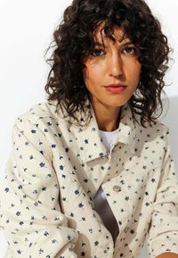 Veste à imprimé floral crème avec des accents bleu marine, dotée d'un col, d'une fermeture à boutons et d'un tissu texturé. Portée par-dessus une chemise blanche.