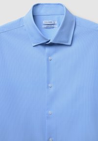 Camicia azzurra chiara, facile da stirare, con una finitura testurizzata. Presenta un colletto a punta e cinque bottoni bianchi lungo la parte anteriore. Etichetta del marchio visibile.